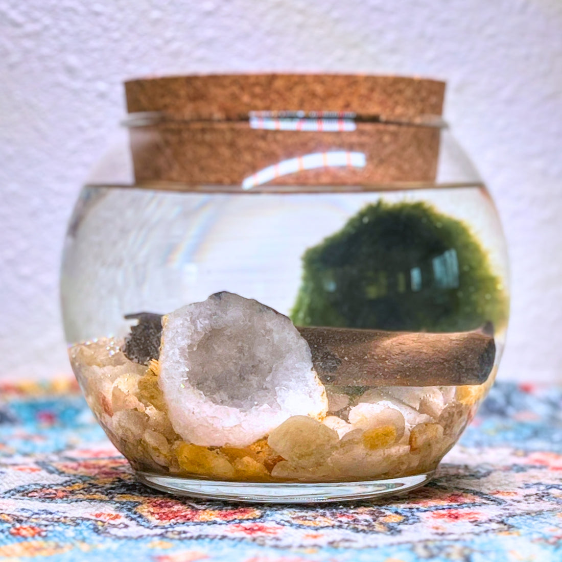 A round glass aquarium with a cork lid on a multicolored textile surface. It contains a large geode, small polished river stones,