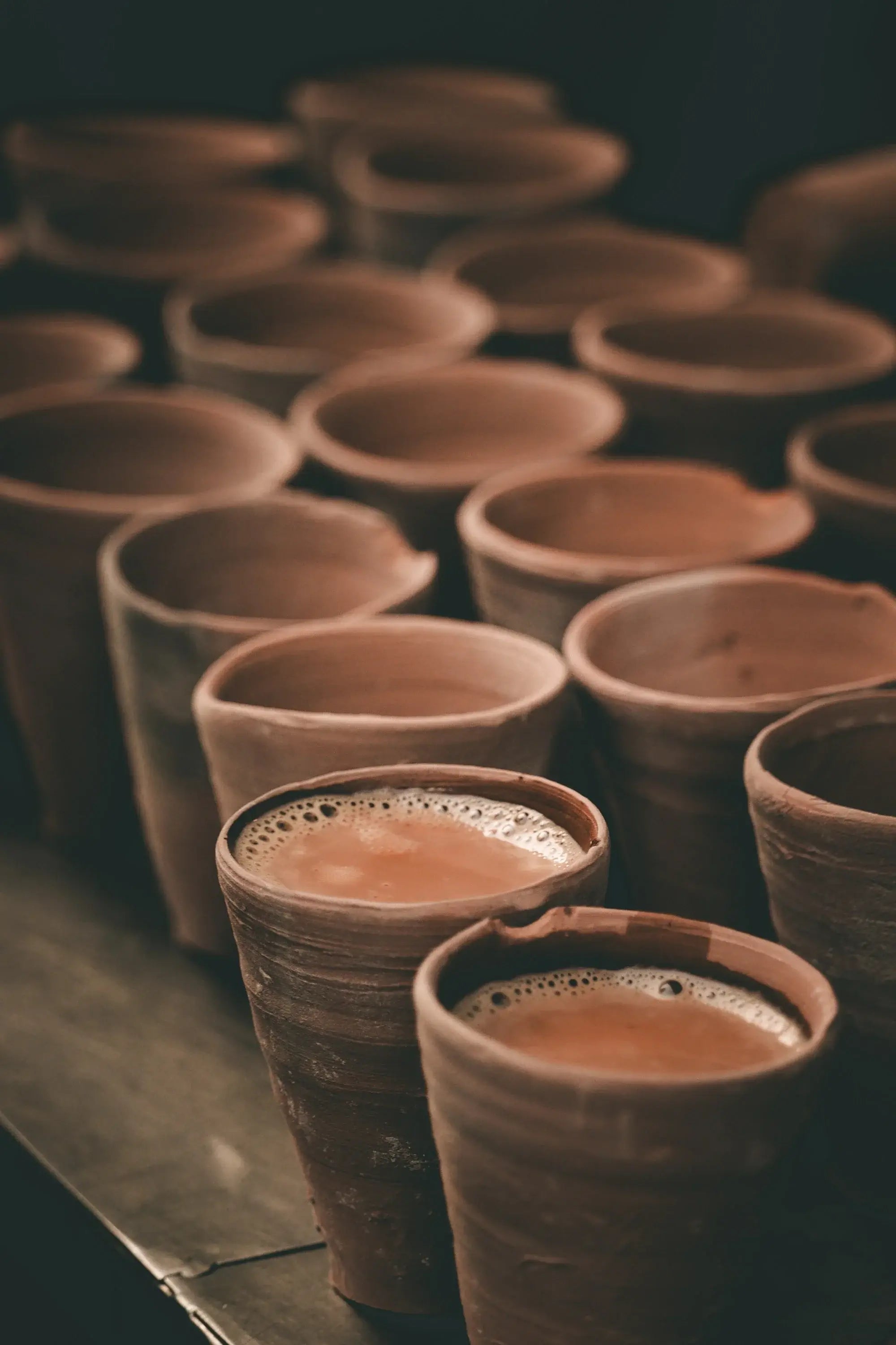 Multiple clay cups with Sacred Plant Co Tulsi Chai Tea, showcasing premium quality herbs, set against a blurred background, with angled perspective, warm earthy tones, and natural clay texture.