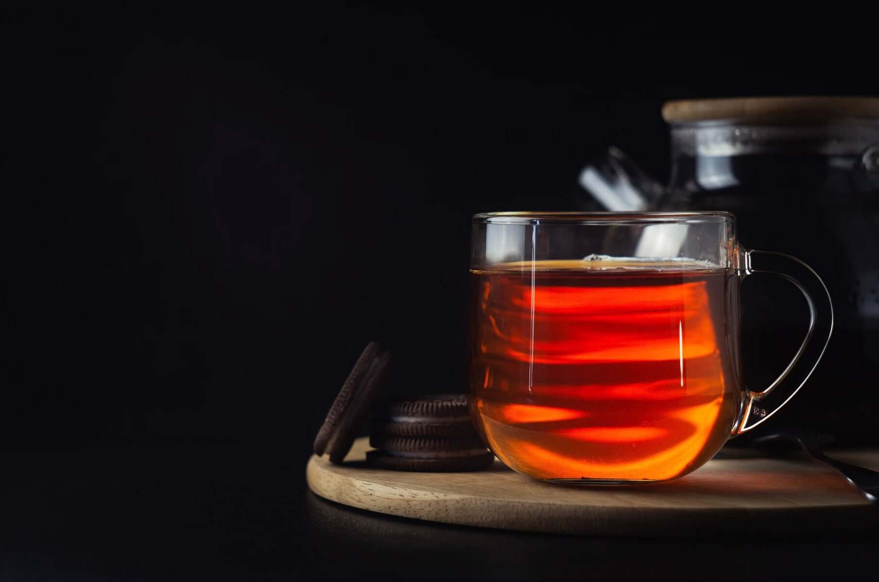 Dark, moody image of glass teacup with Sacred Plant Co Floating Guru Yogi Tea, reflecting warm colors, beside chocolate biscuits on a light wood surface.