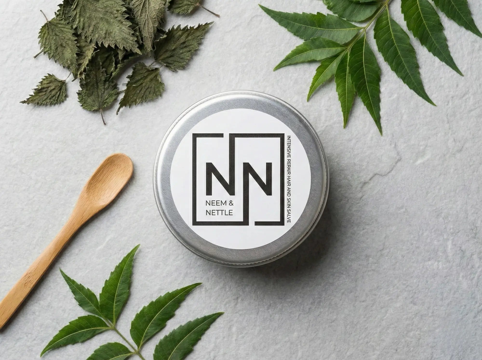 Neem & Nettle salve tin labeled "NN" arranged on a grey surface with a wooden applicator spoon and dried herbal ingredients.