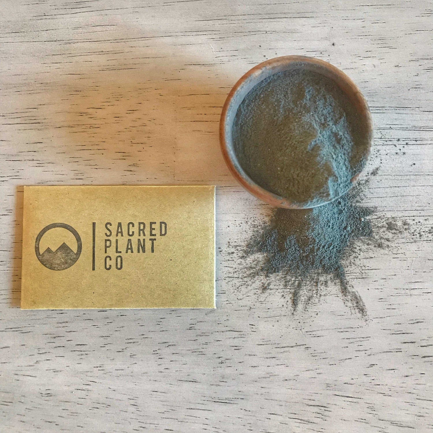 Golden business card for Sacred Plant Co lies next to a wooden bowl of their Ayurvedic face mask powder on a white wooden table, highlighting the natural, earthy ingredients of the product.Plant Co is partially covered by the leaves, and a small wooden bowl rests in the background.