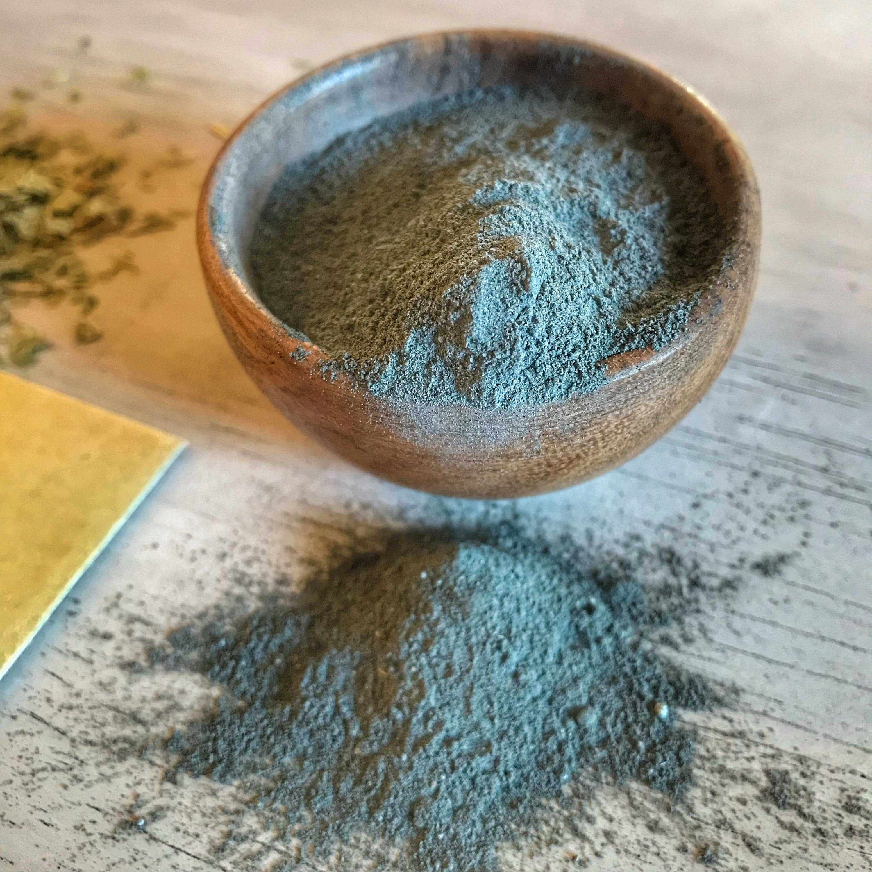 A wooden bowl filled with fine, greenish-gray Ayurvedic face mask powder on a white wooden background, with some of the powder spilled onto the surface, highlighting the natural texture and color of the product.
