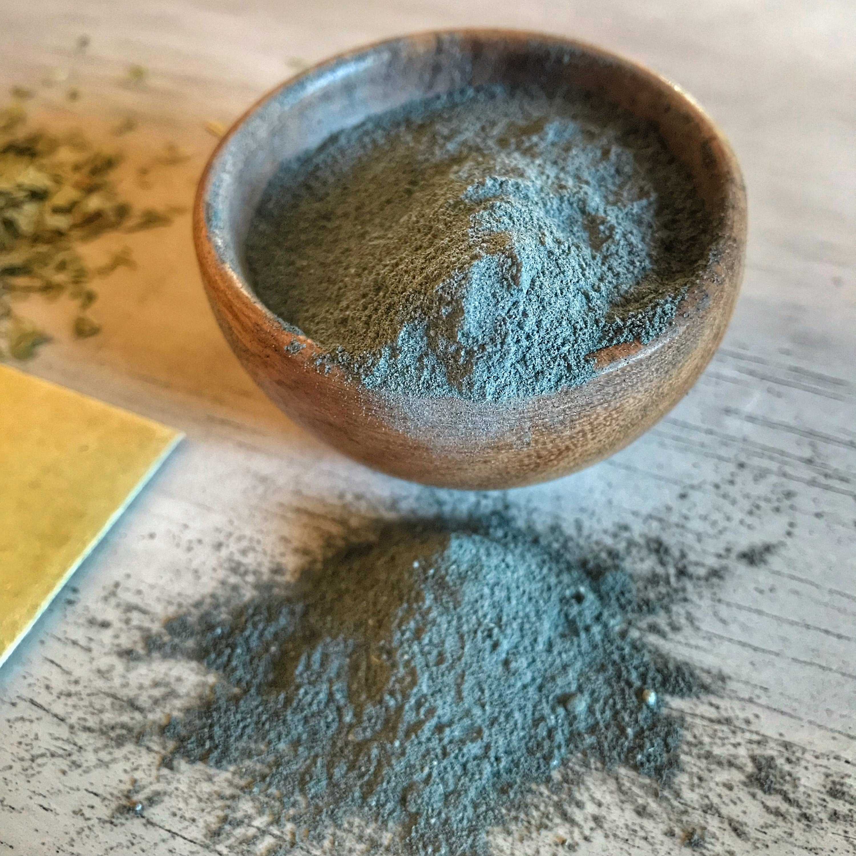 A wooden bowl filled with fine, greenish-gray Ayurvedic face mask powder on a white wooden background, with some of the powder spilled onto the surface, highlighting the natural texture and color of the product.