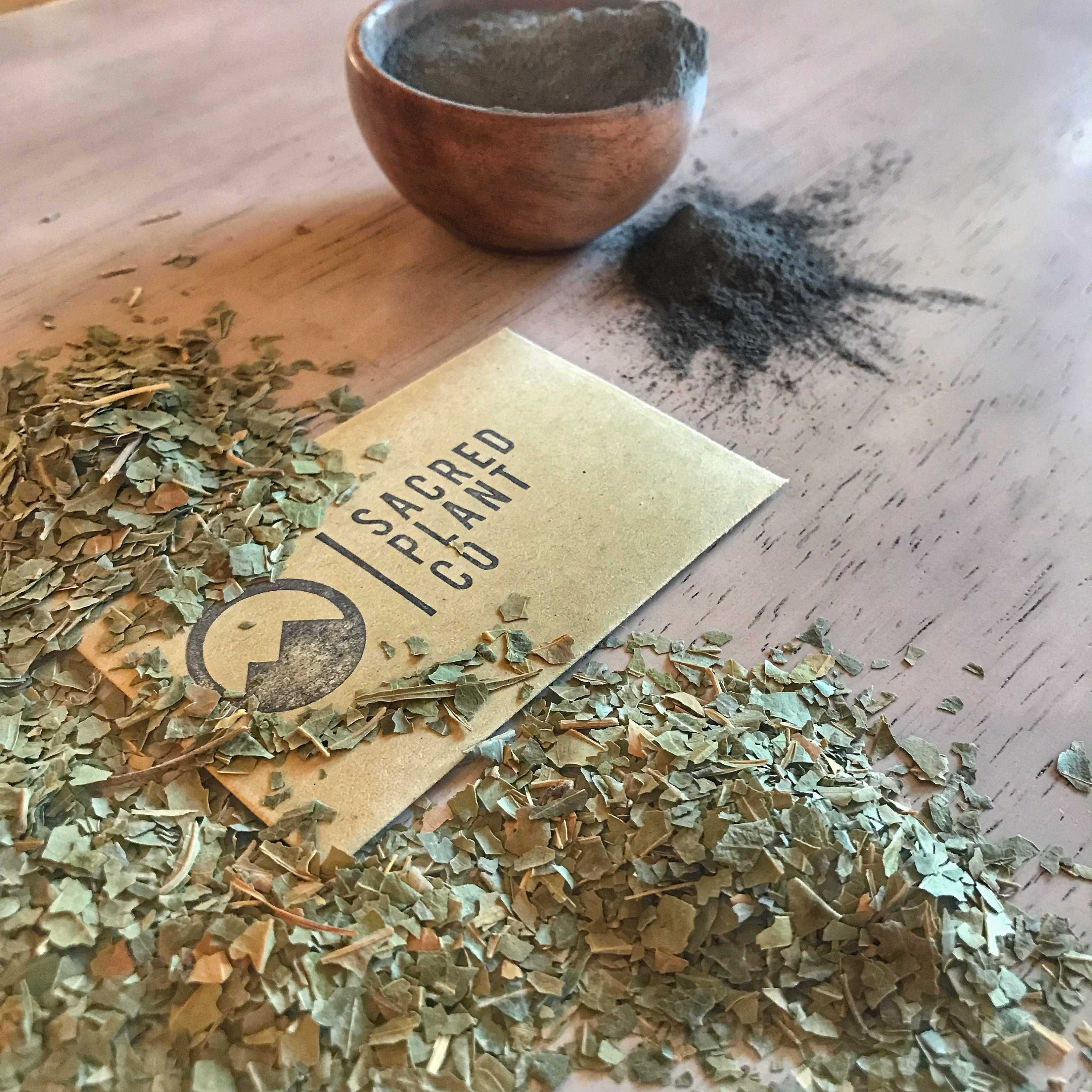 A dynamic arrangement on a wooden surface, featuring a scattering of crushed green leaves and a sprinkling of gray face mask powder. A golden business card for Sacred Plant Co is partially covered by the leaves, and a small wooden bowl rests in the background.