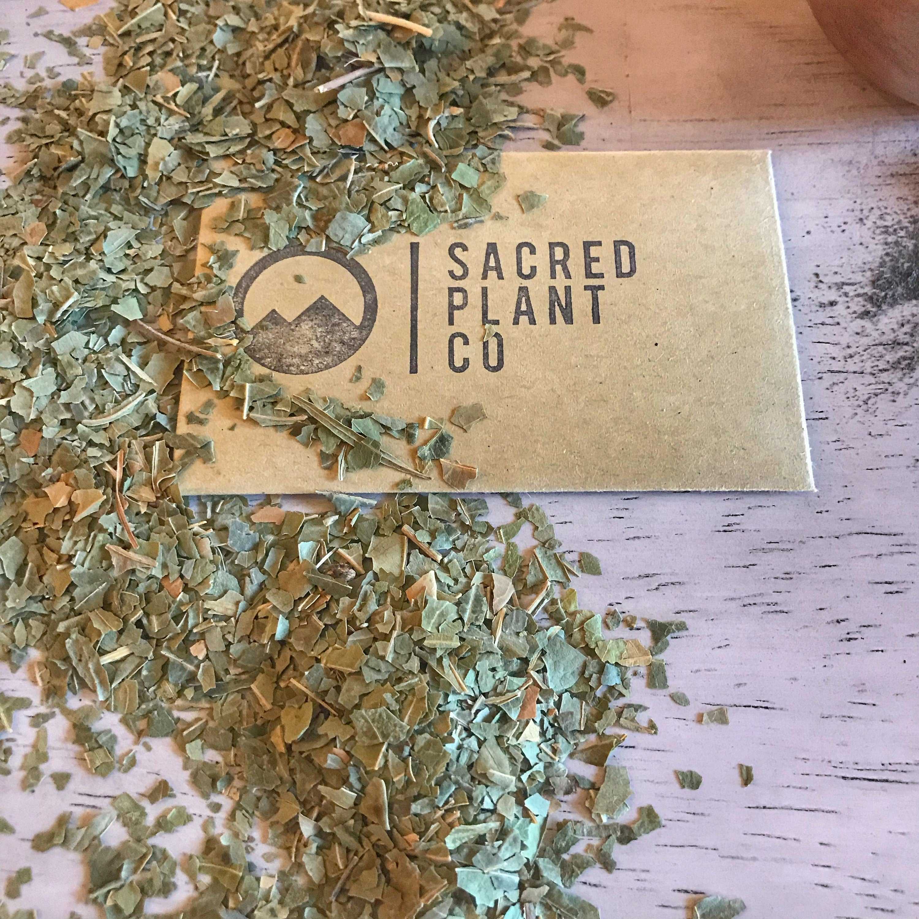 A close-up of dried neem leaves scattered across a white wooden surface, partially covering a golden business card for Sacred Plant Co with their mountain logo, showcasing the natural ingredients used in their products.uthentic Ayurvedic Facial Care Face &amp; Body All Sacred Plant Co