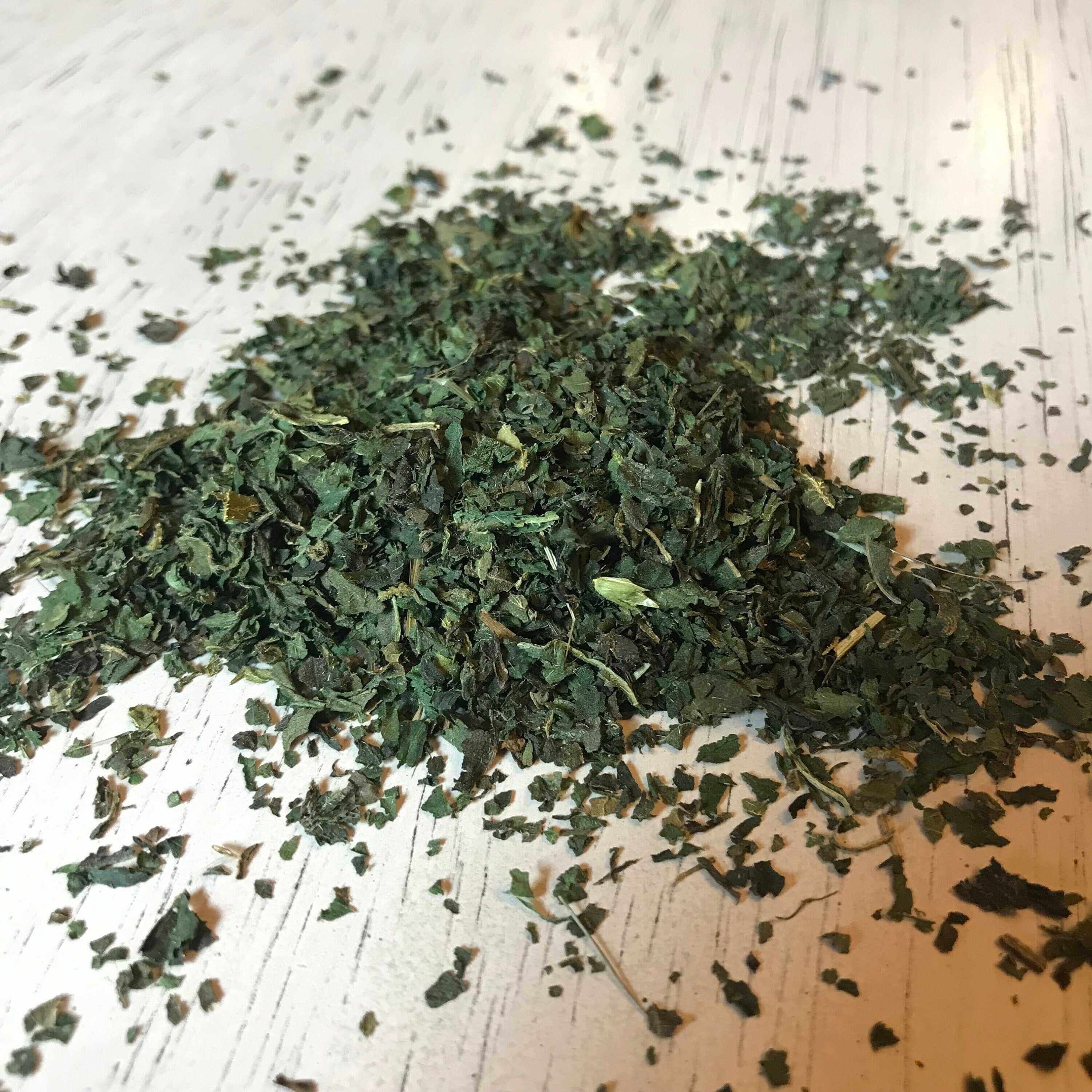 A heap of dried Stinging Nettle Leaves spread across a white wooden surface. The leaves are finely cut and feature a variety of green shades, indicating their natural state. Textures of the wooden surface add to the organic feel of the image, highlighting the nettle&