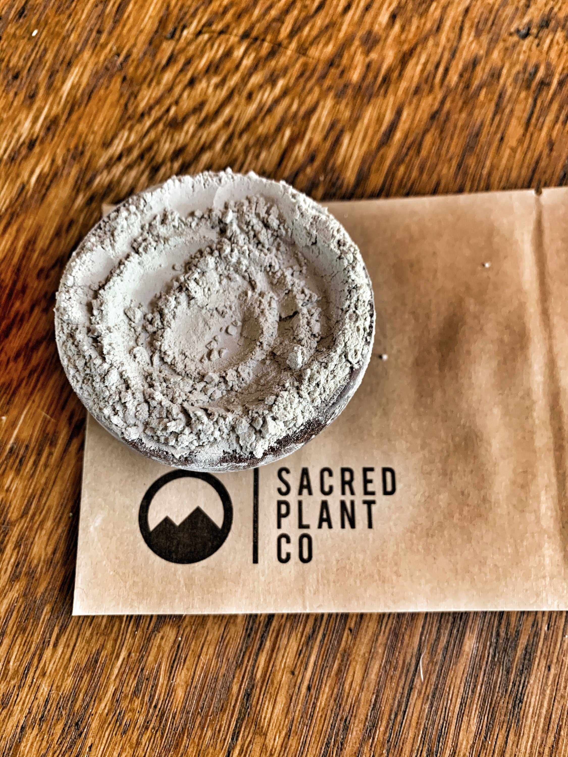 Earthy textured, high-quality Bentonite Clay Powder in a wooden bowl, with Sacred Plant Co kraft paper, shot from an overhead angle.