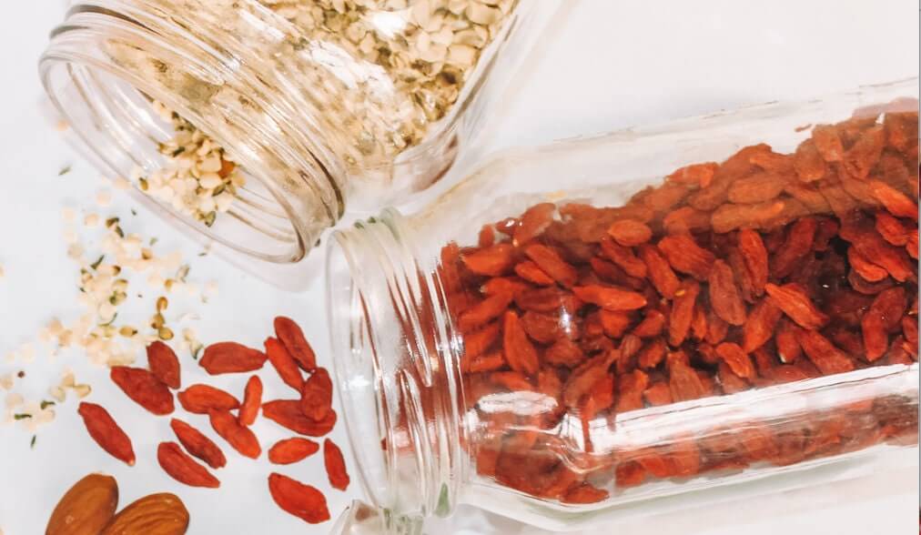 Glass jars spilling premium dried goji berries and hemp seeds onto a white surface. Vibrant red goji berries contrast with creamy hemp seeds, showcasing their natural textures. Perfect for smoothies, snacks, and wellness blends.