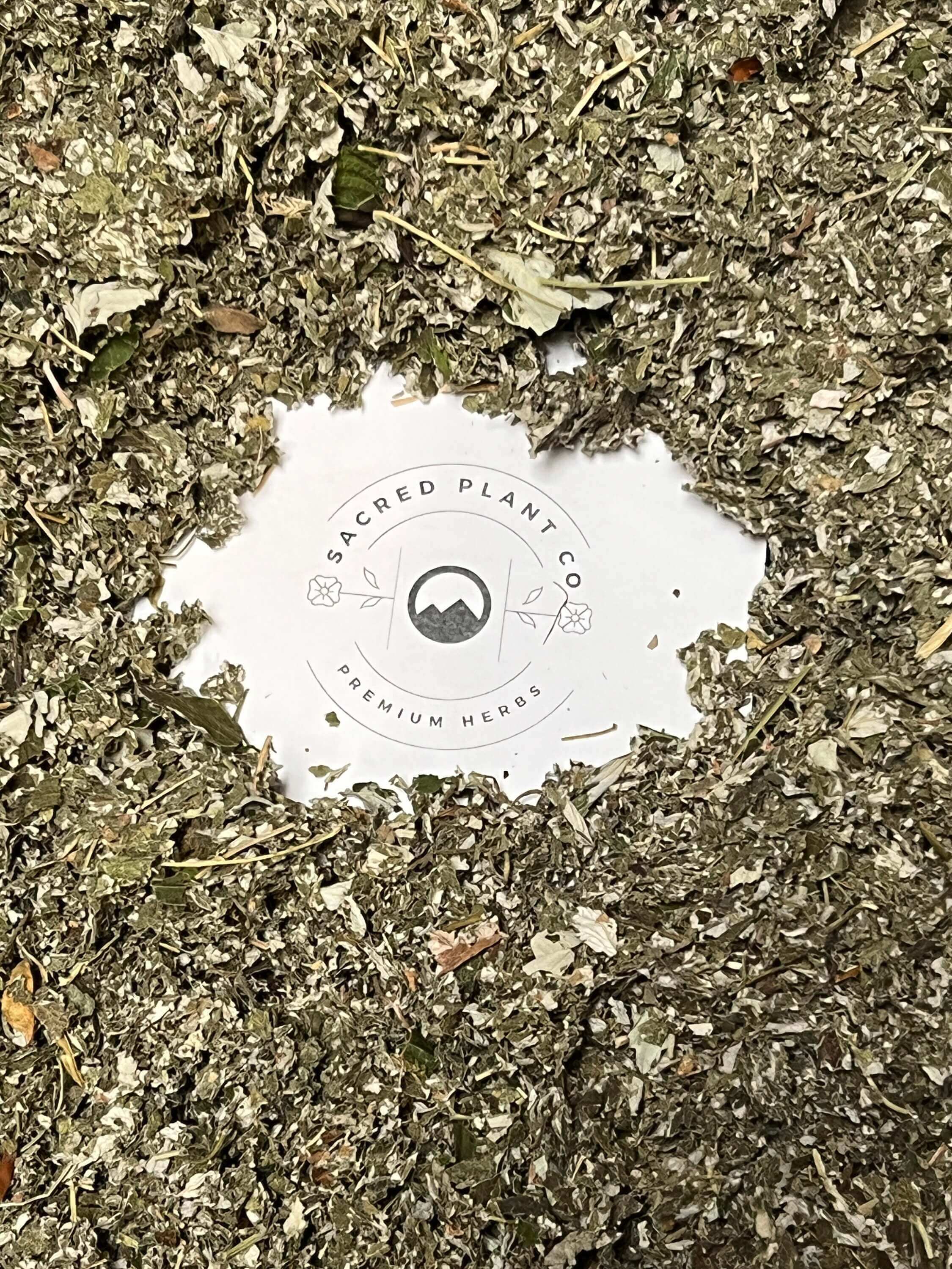 A close-up of loose dried Raspberry Leaf herbs spread out, with the round Sacred Plant Co logo in the center showing &