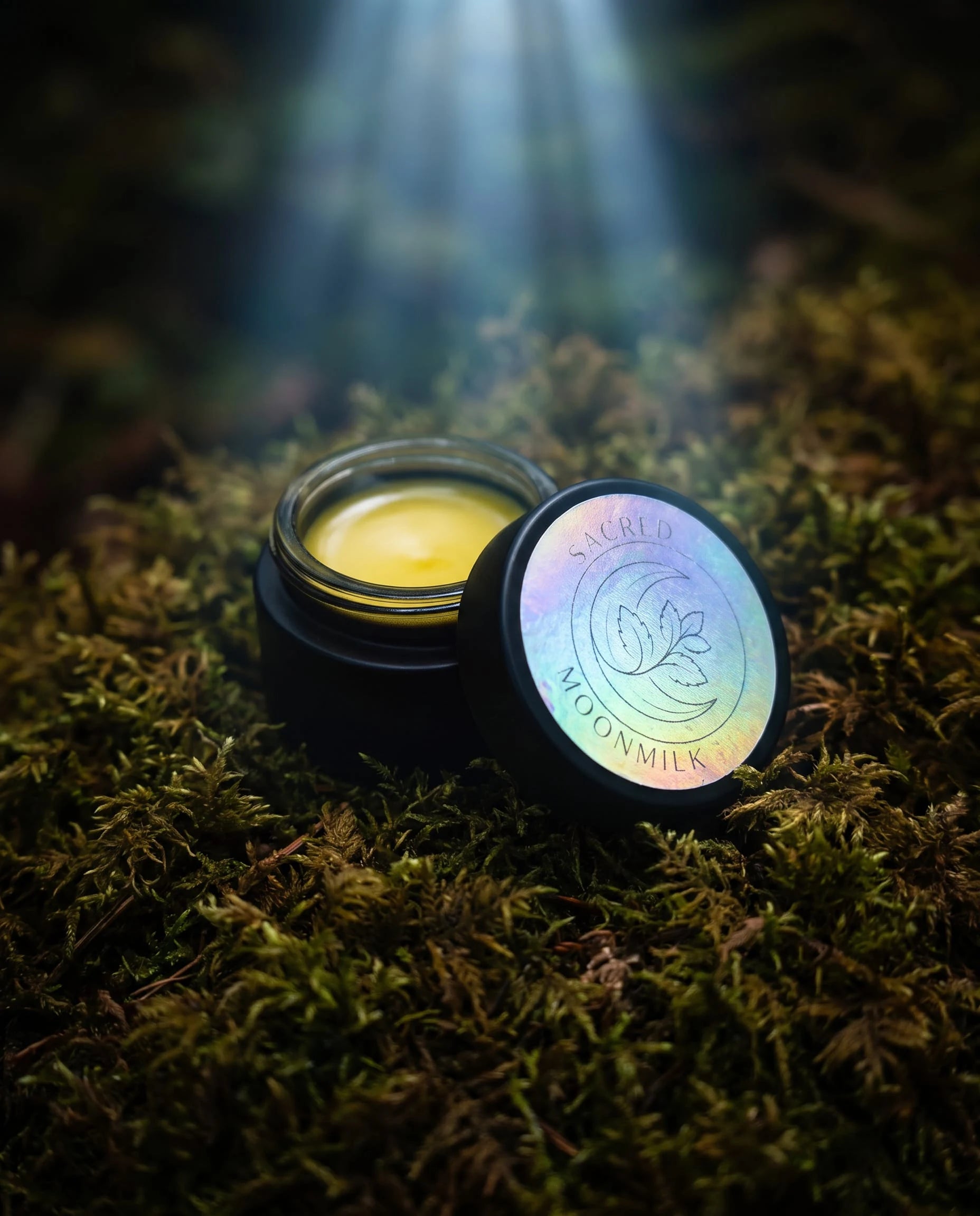 Close-up of an open Moonmilk Botanical Night Balm jar showing the creamy golden balm and the holographic Sacred Moonmilk moon-and-botanical lid on moss