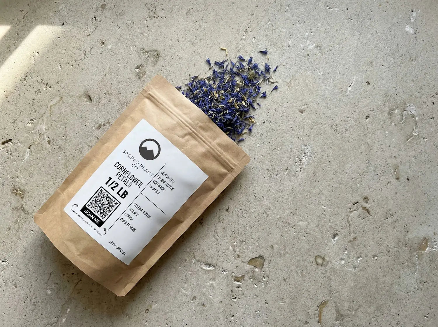 Sacred Plant Co Cornflower Petals 1/2 LB kraft paper bag lying flat on a textured stone surface, with dried blue petals spilling naturally from the top
