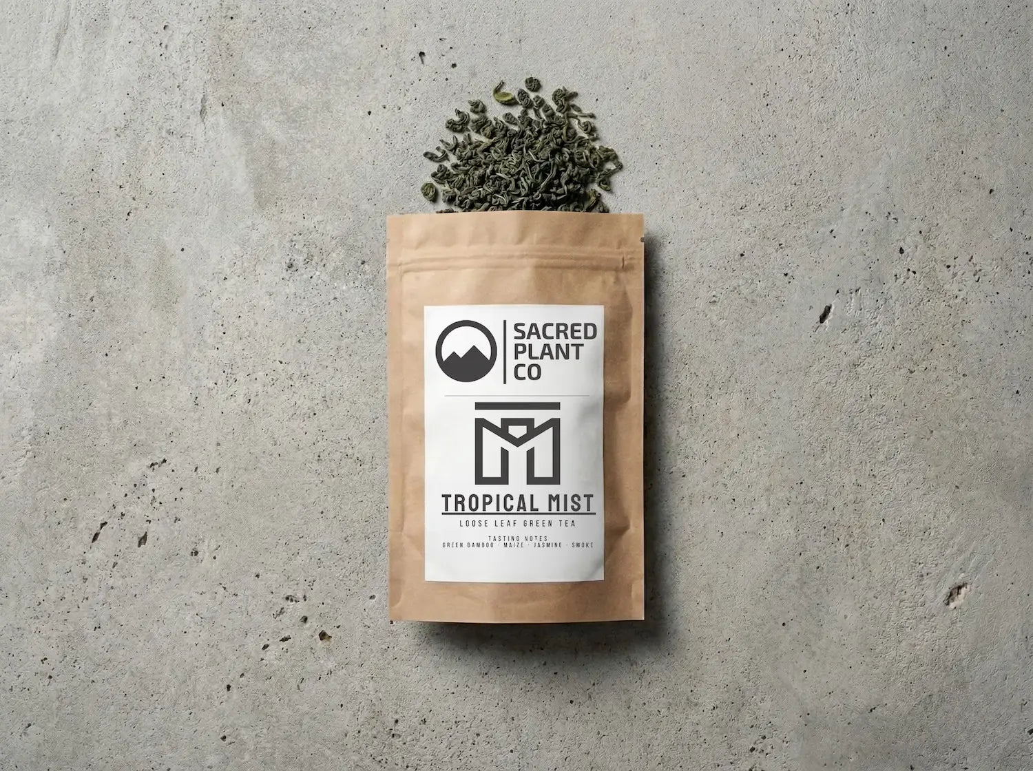 Sacred Plant Co. Tropical Mist loose leaf green tea in a brown kraft bag, layflat editorial photography on a textured stone surface. Premium Chun Mei style green tea leaves with tasting notes of green bamboo, maize, jasmine, and smoke spilling naturally from the packaging.