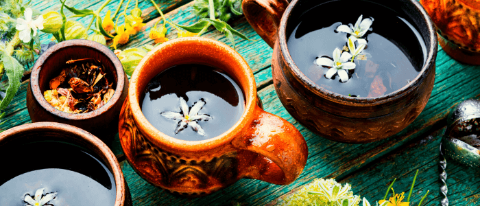 Three brown ceramic cups of Slide Rock Bolter Wild Colorado Tea sit on a teal blue wooden table, each with a unique floral garnish. A bowl of dried herbs and flowers accompanies them, set against a backdrop of scattered yellow and green botanicals, invoking the natural essence of Colorado&