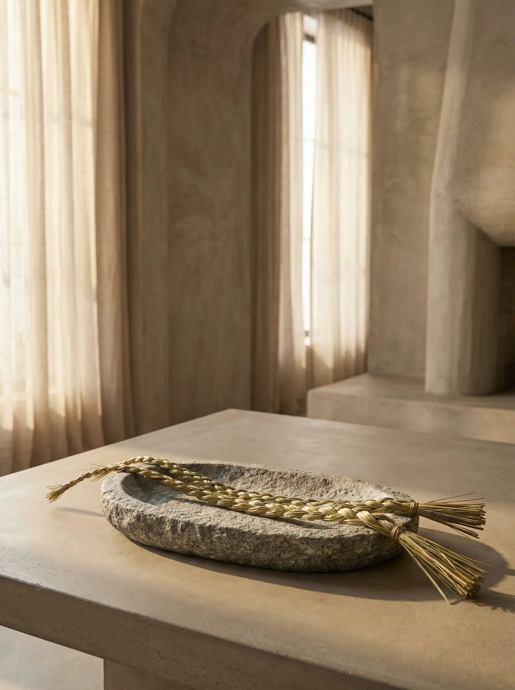 Long braided sweetgrass displayed in a stone tray inside a minimalist brutalist home with soft filtered sunlight.