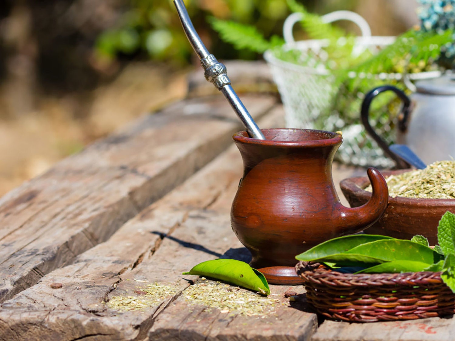 Traditional yerba mate in a brown gourd with a metal bombilla, on a rustic wooden table with loose mate leaves and greenery.