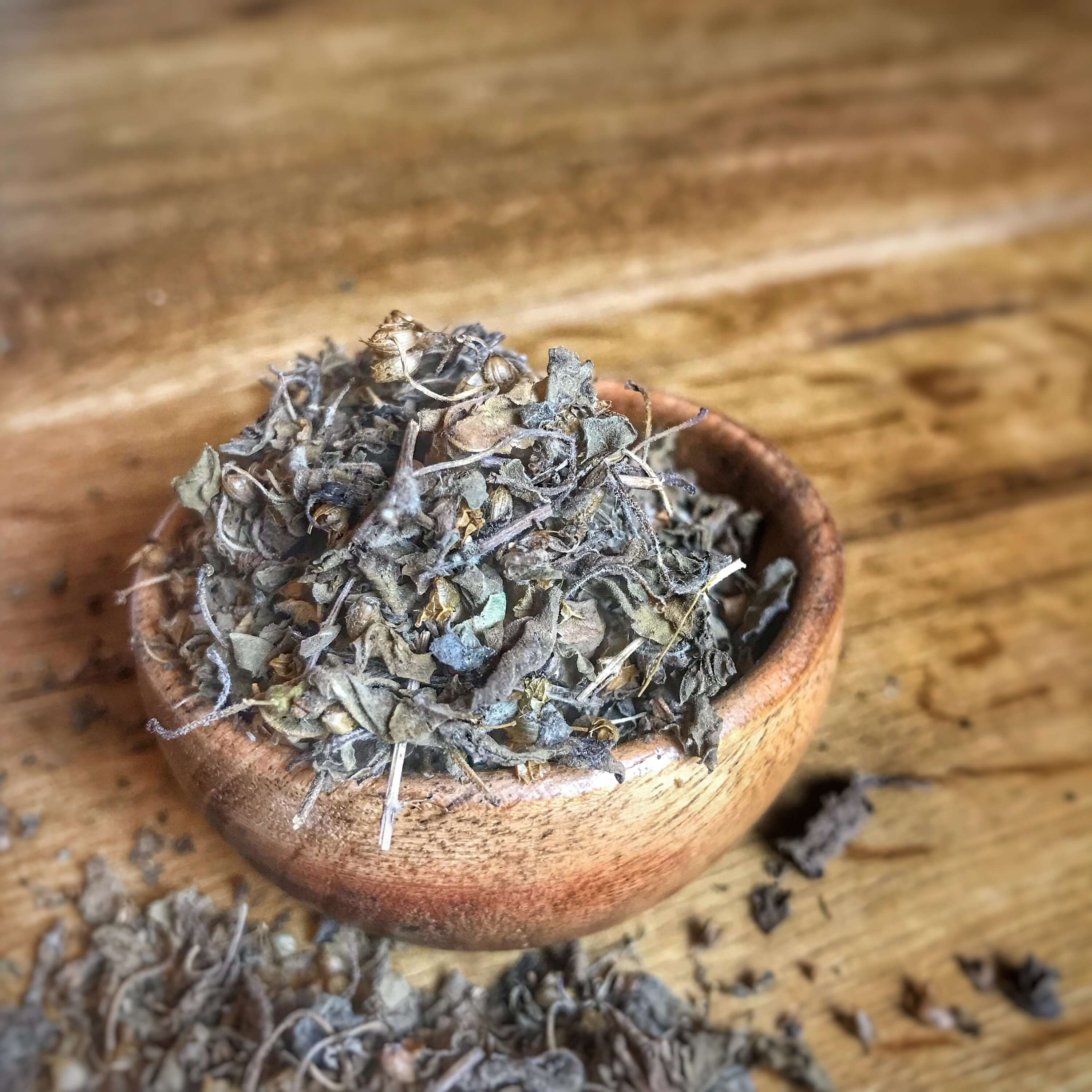 Sacred Plant Co close up image of organic dried Tulsi Herbs spilling from a wooden bowl, highlighting the natural texture and rustic wooden setting.