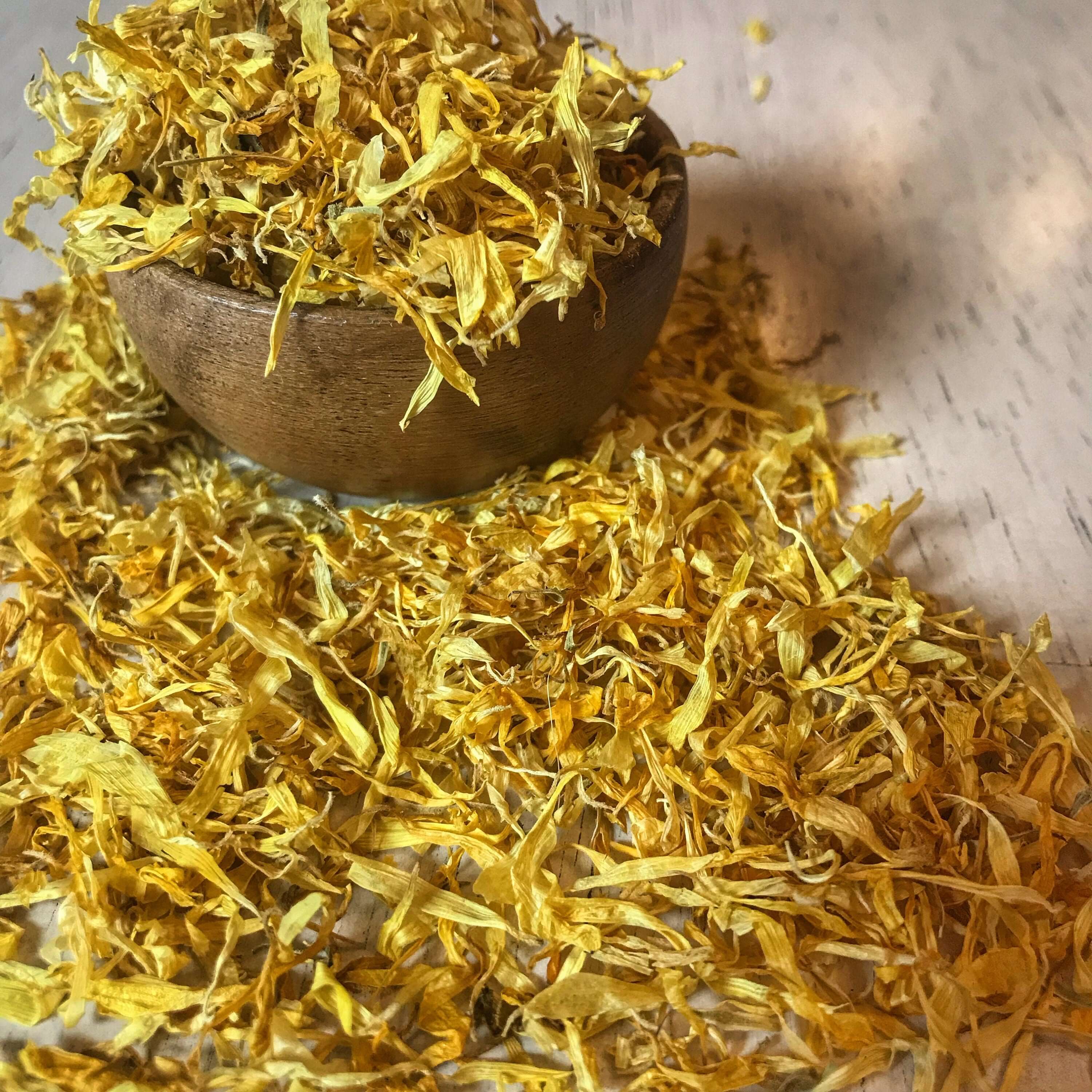 Sacred Plant Co premium Calendula Petals in a rustic bowl on a pale surface, with a sunlit backdrop accentuating their vibrant yellow hues.
