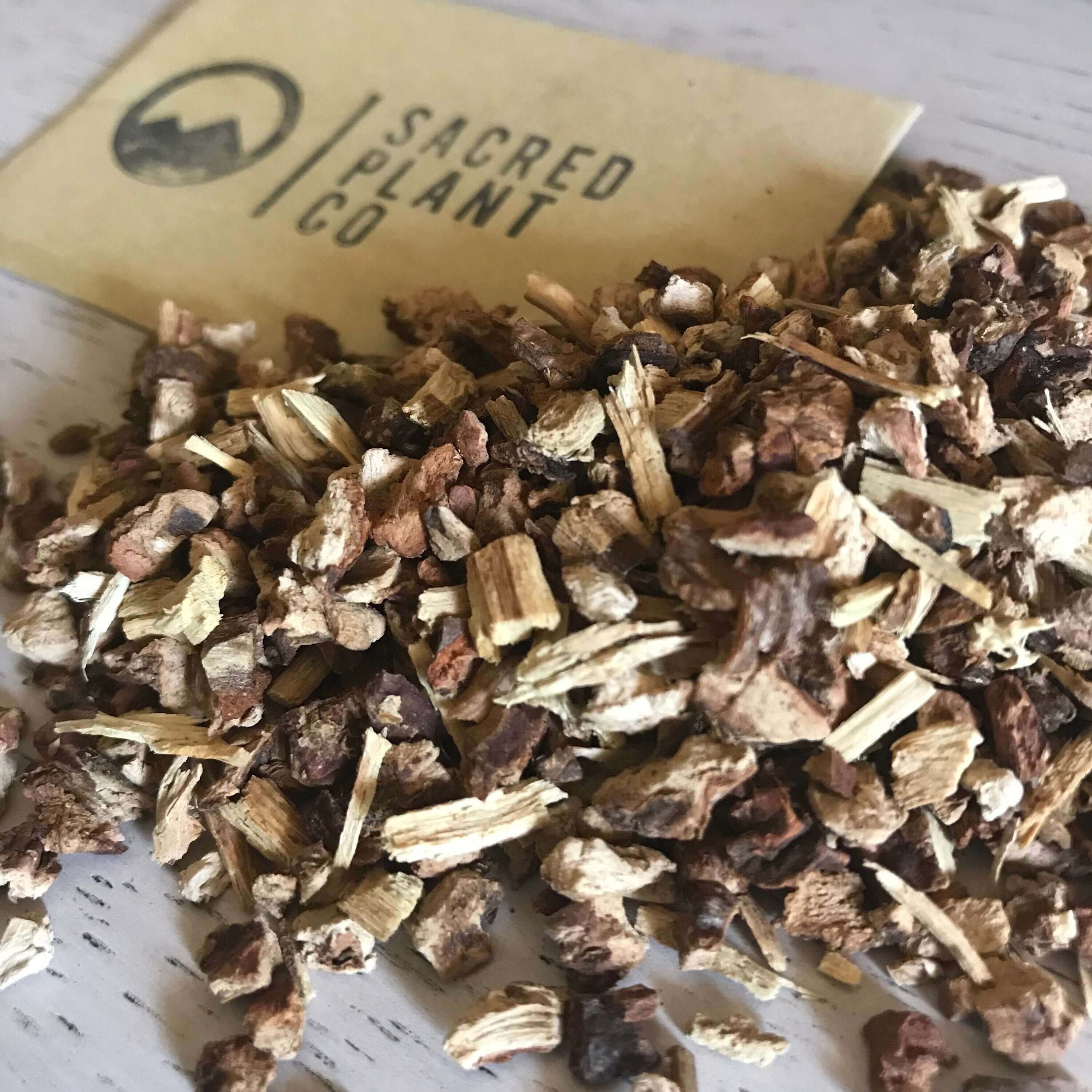Coarsely chopped Sarsaparilla Root from Sacred Plant Co, featuring natural beige and dark brown pieces with a rough texture on a light wooden surface.