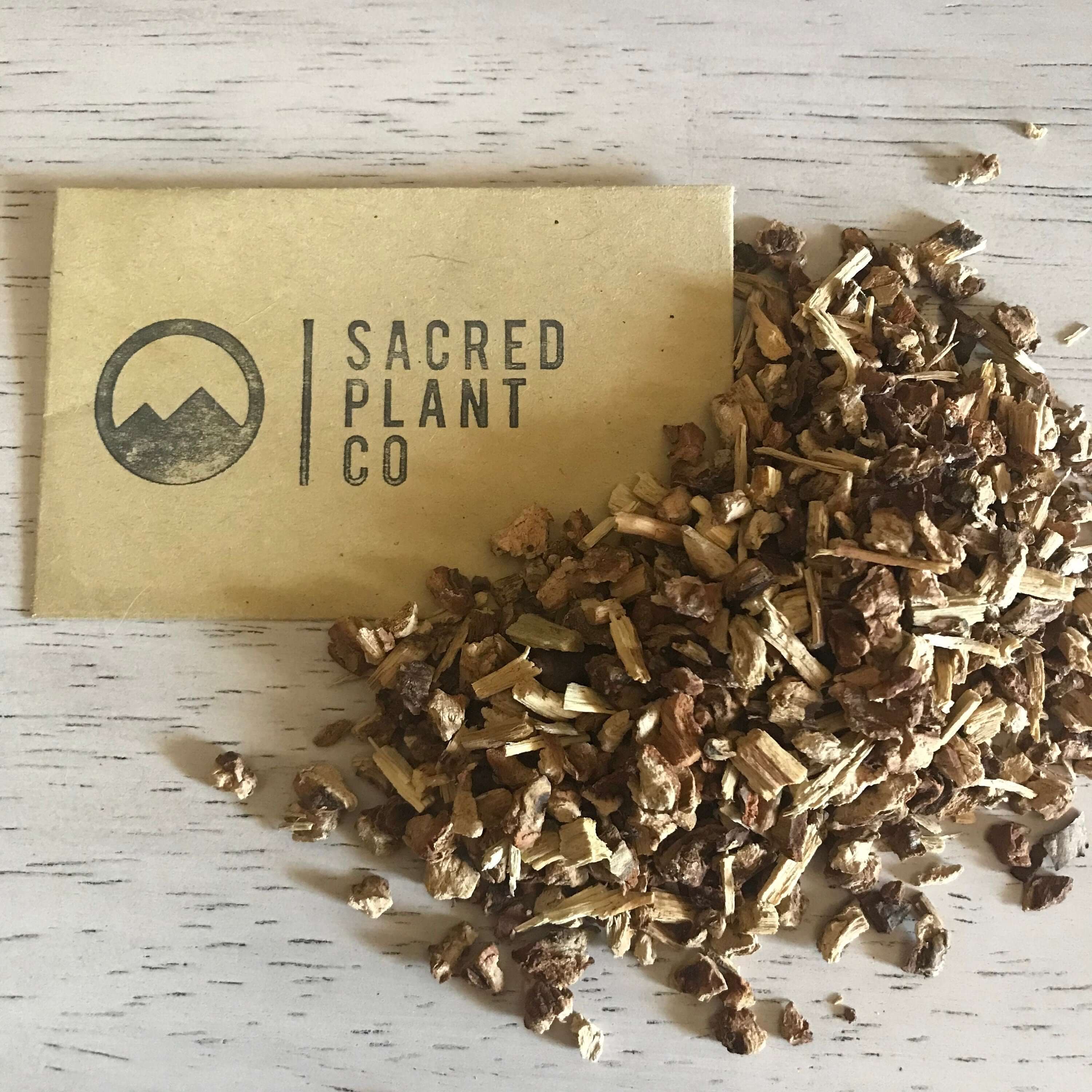 Earthy, dried Sarsaparilla Root pieces from Sacred Plant Co, with a coarse texture and brown tones, resting beside branded packaging.