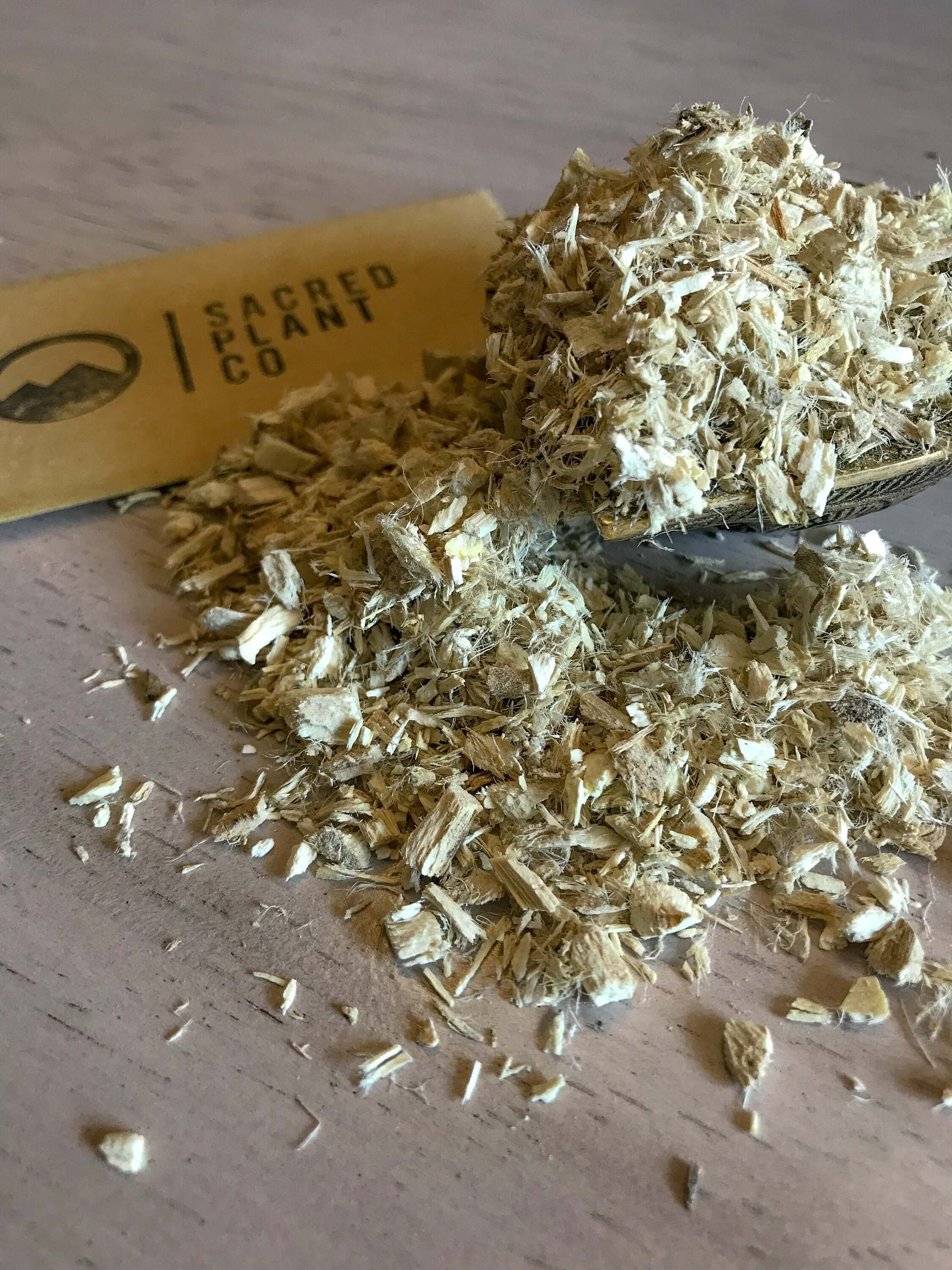 Finely shredded Marshmallow Root in a wooden spoon for oil essence, set against a light wooden table with Sacred Plant Co branding in the background.