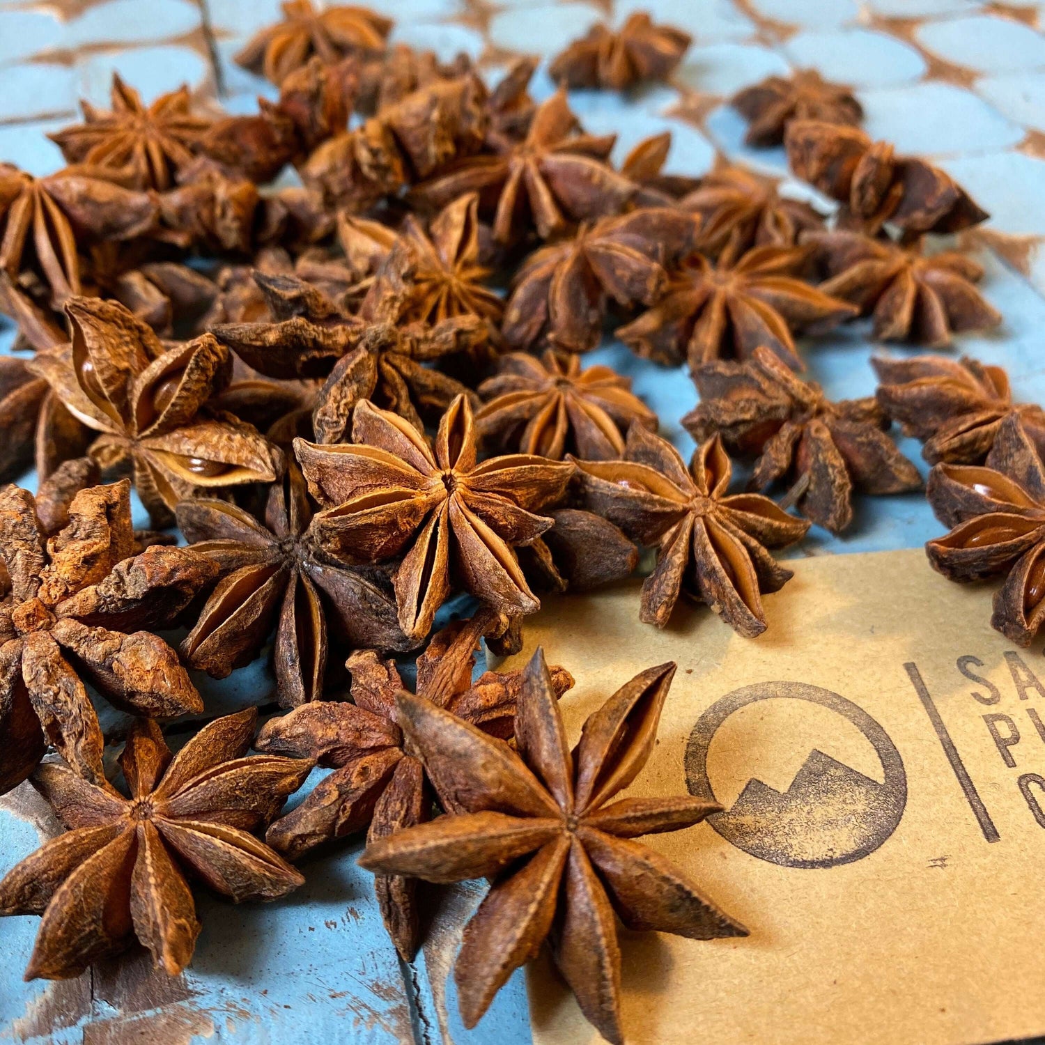 Organic Star Anise pods from Sacred Plant Co, arranged on rustic surface with company branding, capturing dry texture details and aromatic quality on a light blue surface.