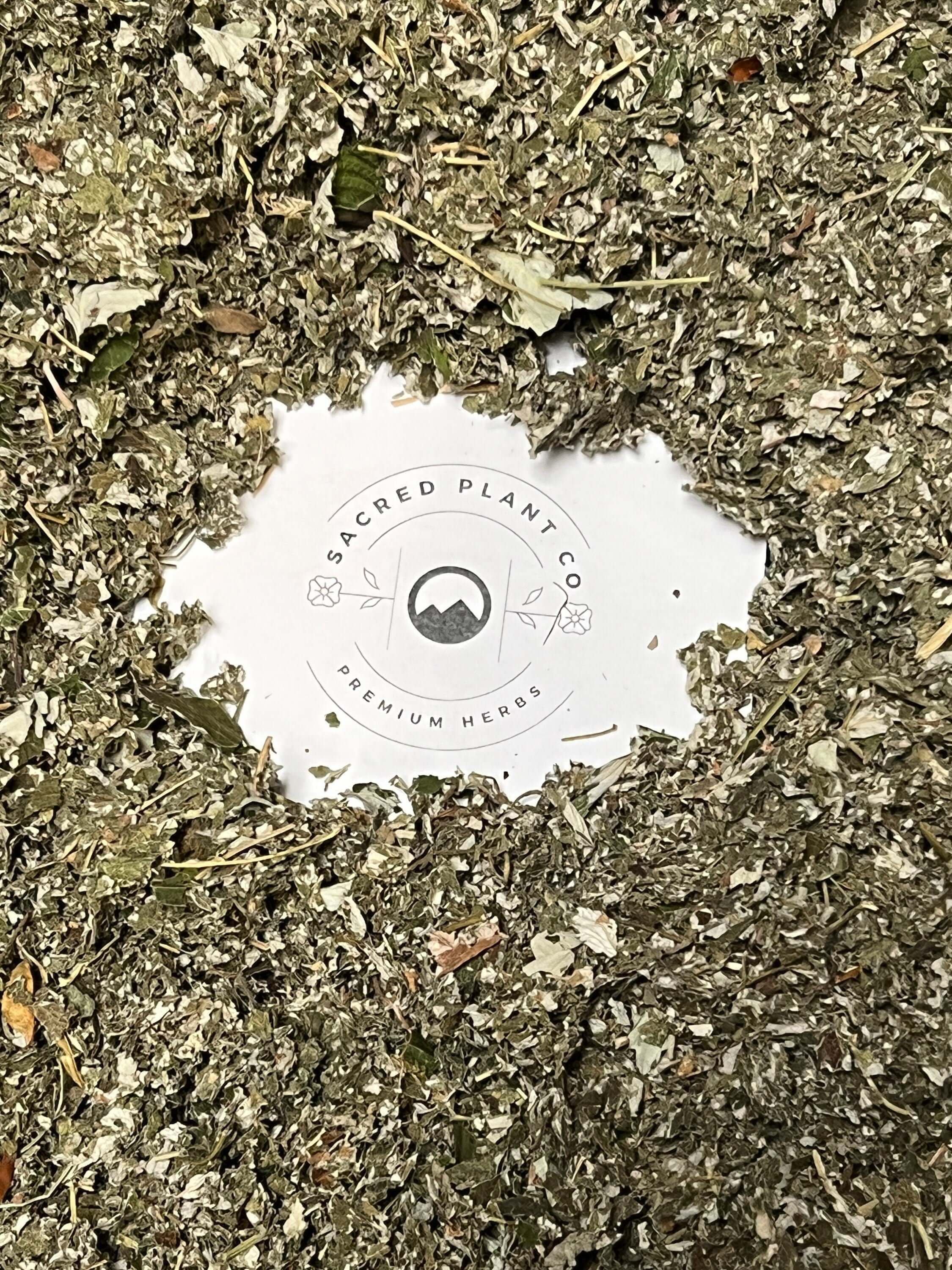 A close-up of loose dried Raspberry Leaf herbs spread out, with the round Sacred Plant Co logo in the center showing &