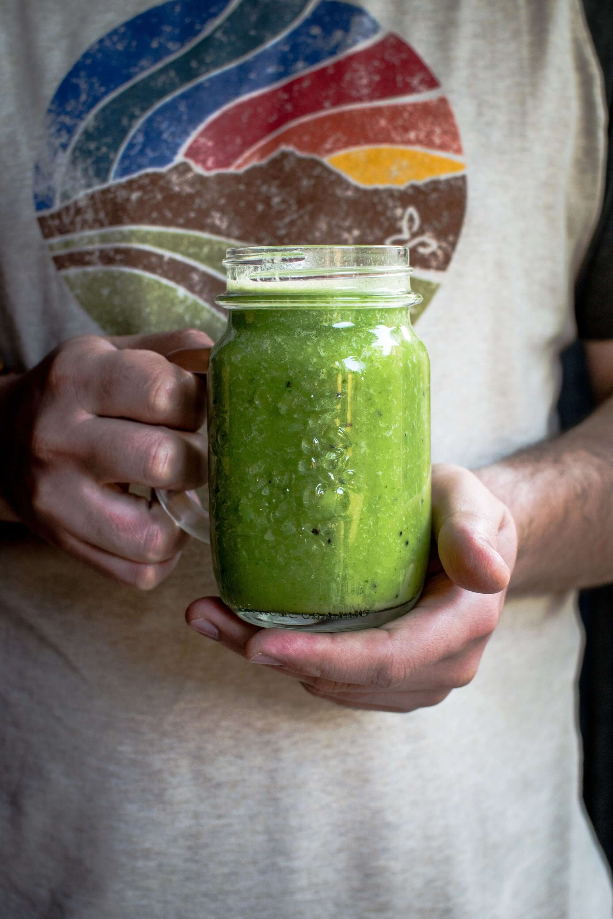 Green Sasquatch Piss Powder smoothie in a mason jar with seeds on top, held by a casually dressed person with simple daily wear, by Sacred Plant Co.