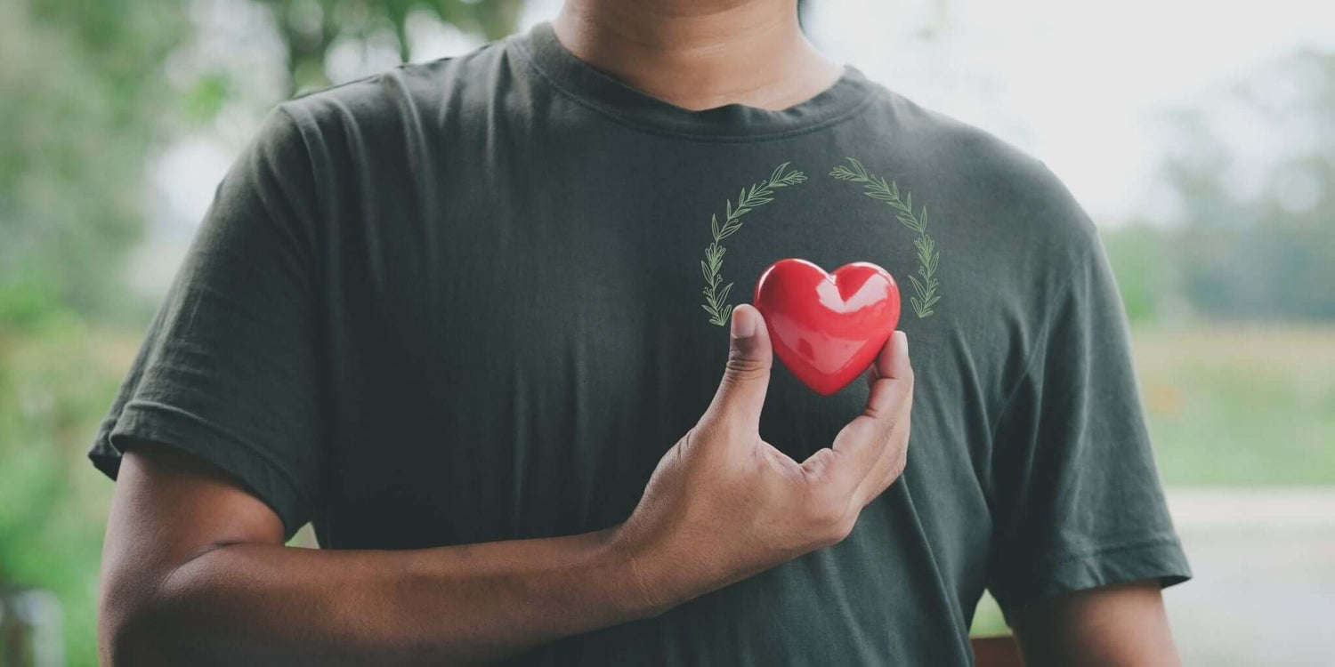 A close-up image of a person wearing a dark green t-shirt, gently holding a bright red heart-shaped object over their chest, symbolizing heart health and care. 