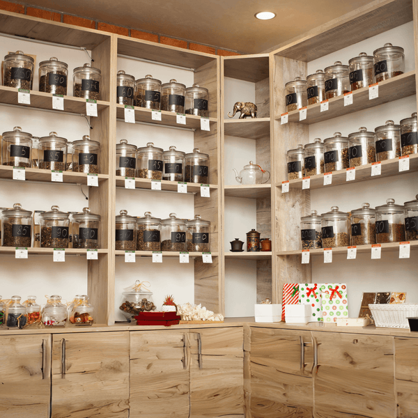 A cozy tea shop interior with natural wood shelves filled with clear glass jars of various loose leaf teas. The shelves are well-stocked with Sacred Plant Co Wholesale Tea, reflecting a warm and inviting ambiance.