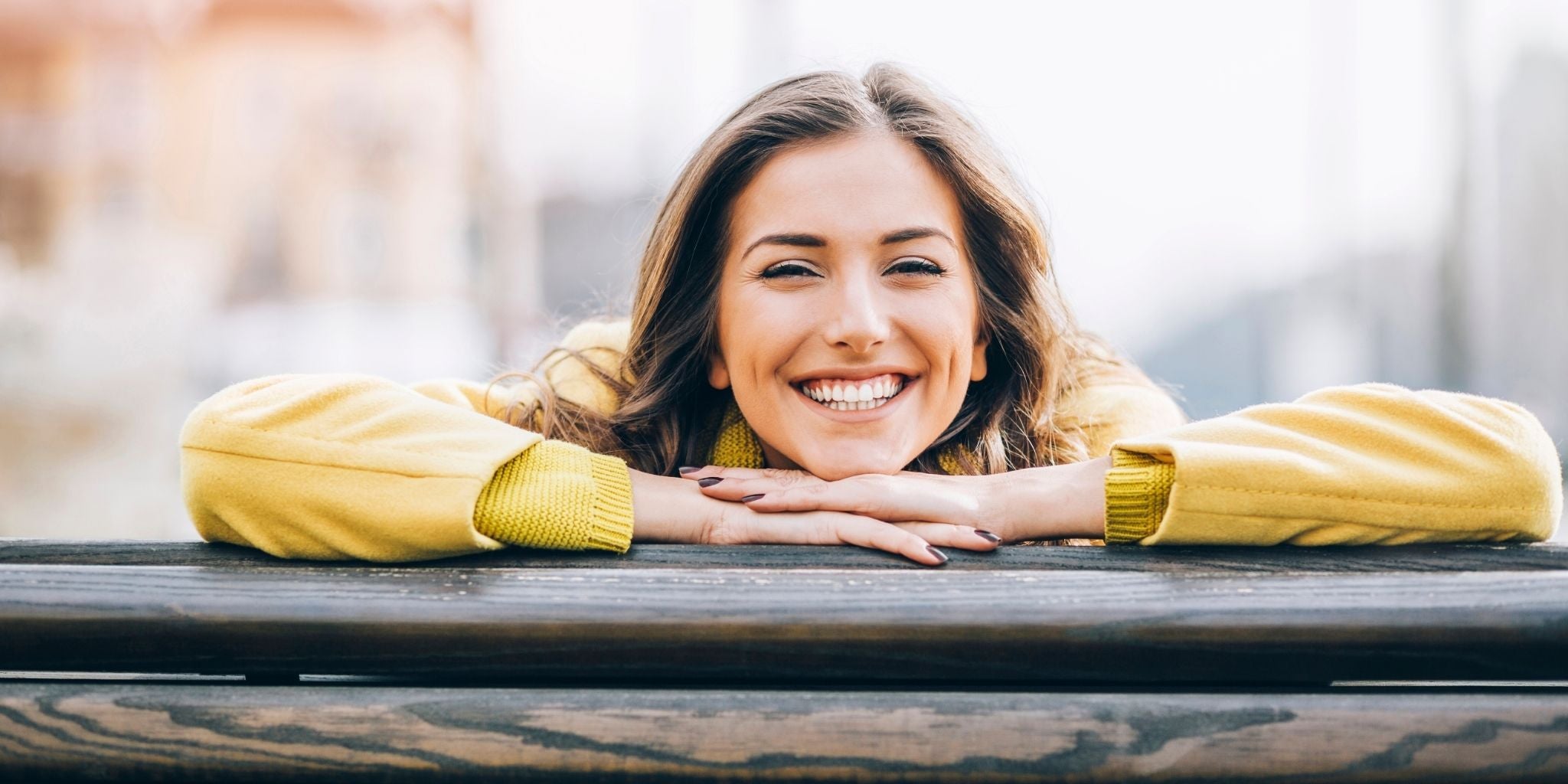 A cheerful woman in a yellow coat leaning on a wooden bench, smiling brightly with a blurred cityscape in the background.