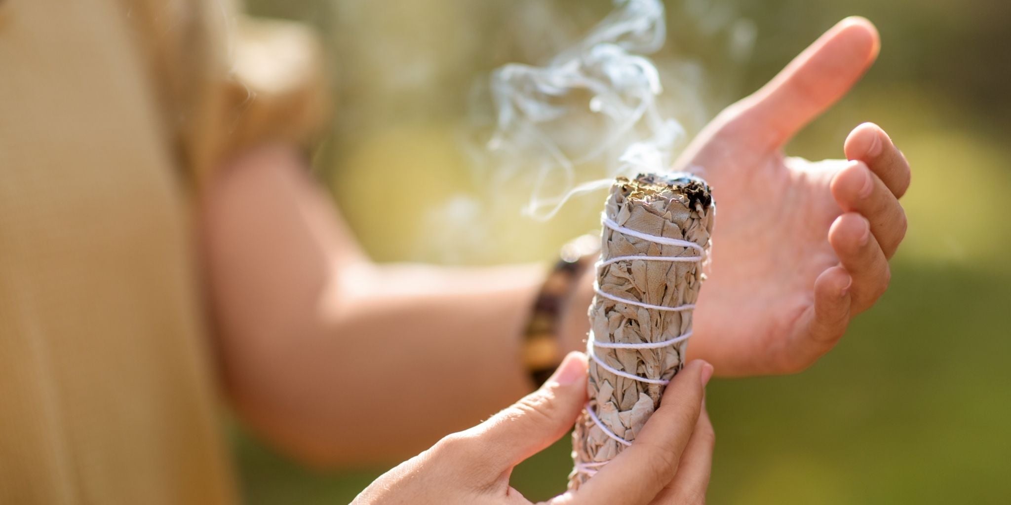 A hand holding a burning sage smudge stick with soft, delicate smoke rising, set against a blurred natural outdoor background.
