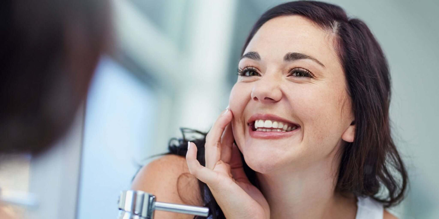 Smiling woman looking at her reflection in a mirror, showcasing clear and radiant skin, representing natural solutions for achieving a healthier complexion.