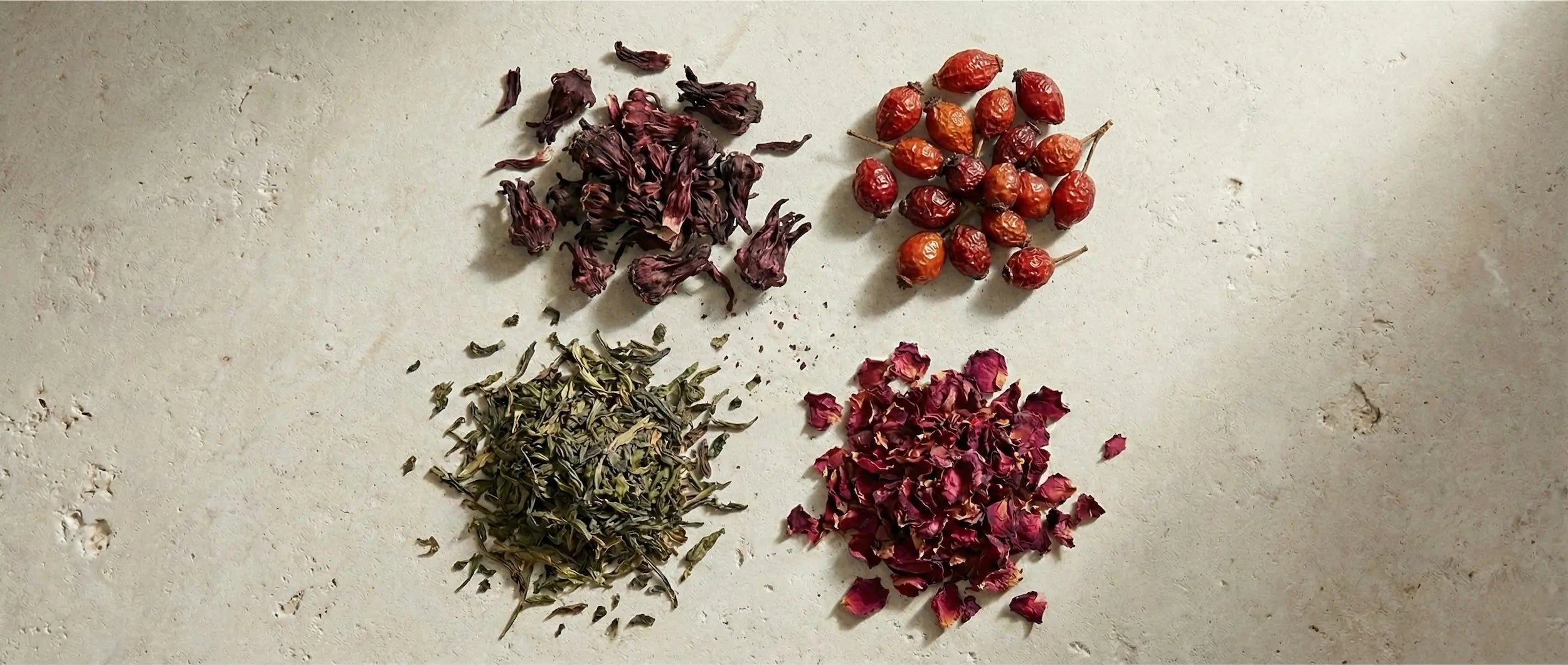 Four distinct piles of dried herbs on a stone surface: deep red hibiscus, whole orange rose hips, green tea leaves, and pink rose petals.