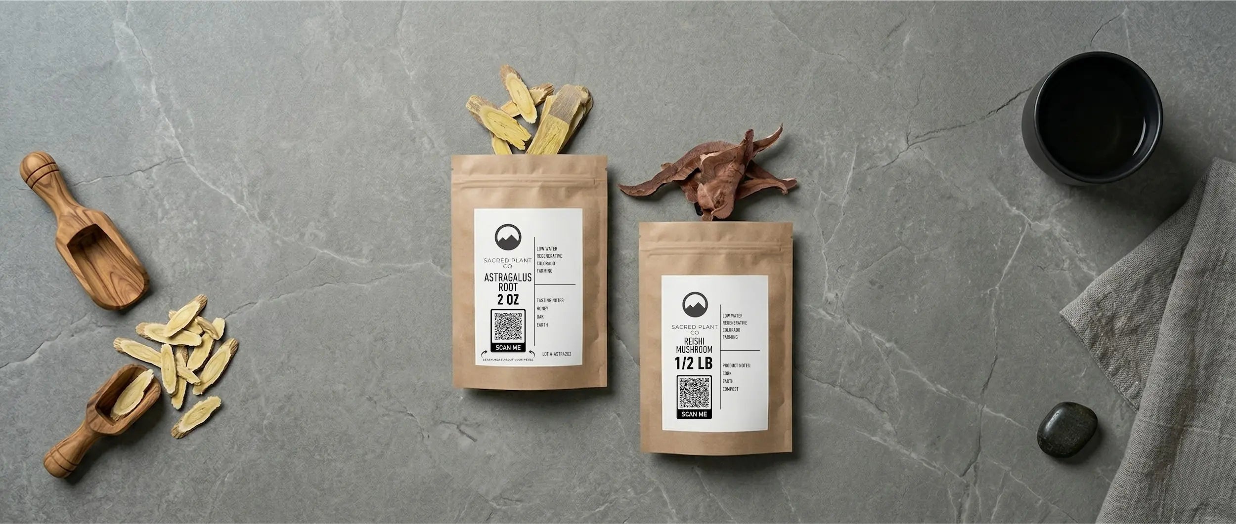Top-down view of Sacred Plant Co whole-slice Astragalus root and Reishi mushroom in kraft packaging on a grey stone surface, accompanied by wooden scoops and a tea cup.