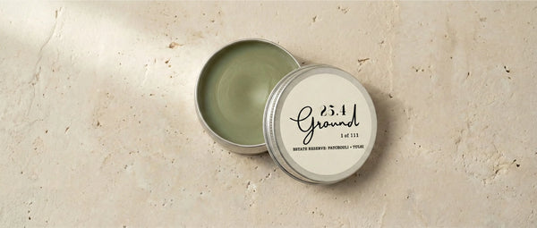 Open tin of 25.4 Ground Estate Reserve balm on travertine stone showing natural green color from cold-infused patchouli and tulsi.
