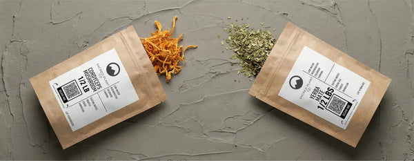 Flat lay of Sacred Plant Co's half-pound kraft bags of bulk Cordyceps Mushroom and Yerba Maté on a textured gray surface. Bright orange dried cordyceps fruiting bodies and crushed green yerba maté leaves spill from the packaging