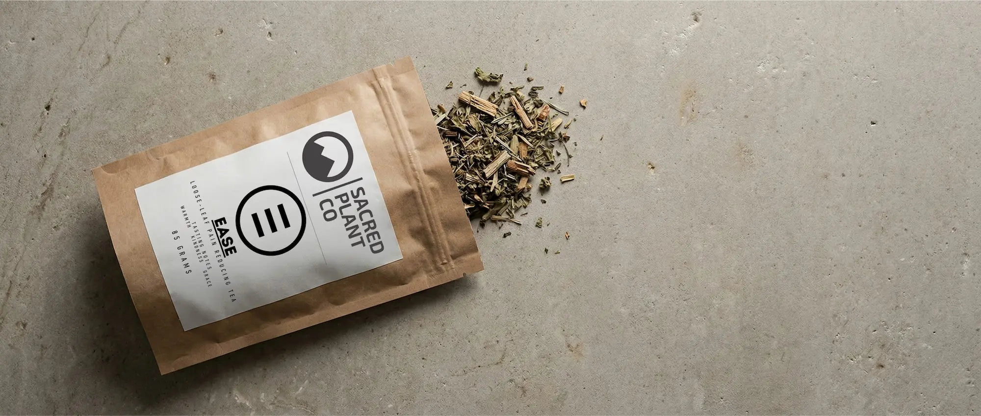 A top-down view of loose-leaf Ease Herbal Tea spilling from a brown kraft paper bag onto a textured stone surface, showing Holy Basil leaves and White Willow Bark pieces.