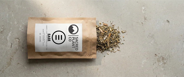 High-quality loose leaf tea spilling from a Sacred Plant Co "Ease" bag onto a stone surface, showing dried herbs and bark.