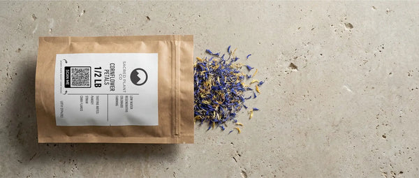 A kraft paper package of Sacred Plant Co cornflower petals spilling vibrant blue and yellow dried herbs onto a stone surface.
