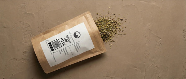 Sacred Plant Co Oregano Leaf 1/2 LB kraft bag on a plaster surface, dried cut and sifted leaf spilling out, QR-coded lot label visible.