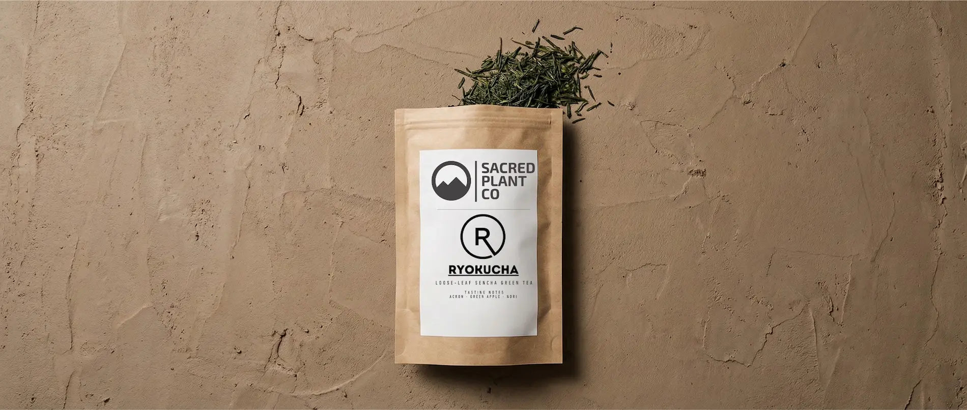 A pouch of Sacred Plant Co Ryokucha Sencha green tea laying on a textured earth surface with dark green, needle-shaped loose leaves spilling out.