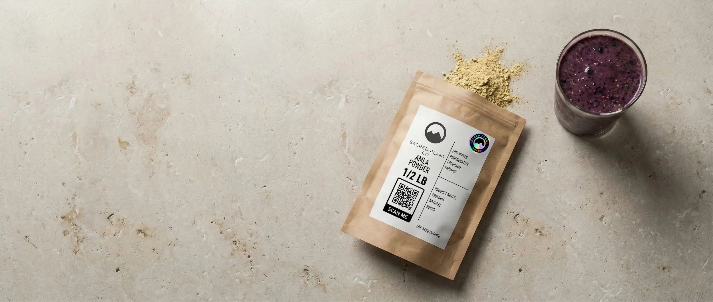 A 1/2 lb bag of Sacred Plant Co Amla Powder sitting on a stone surface next to a finished purple berry smoothie glass.