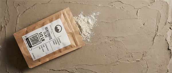 Flat lay of a Sacred Plant Co 1/2 lb Bentonite Clay pouch resting on a textured, wet clay surface. Fine white clay powder spills from the open kraft paper bag, and the label highlights 'Low Water Regenerative Colorado Farming'