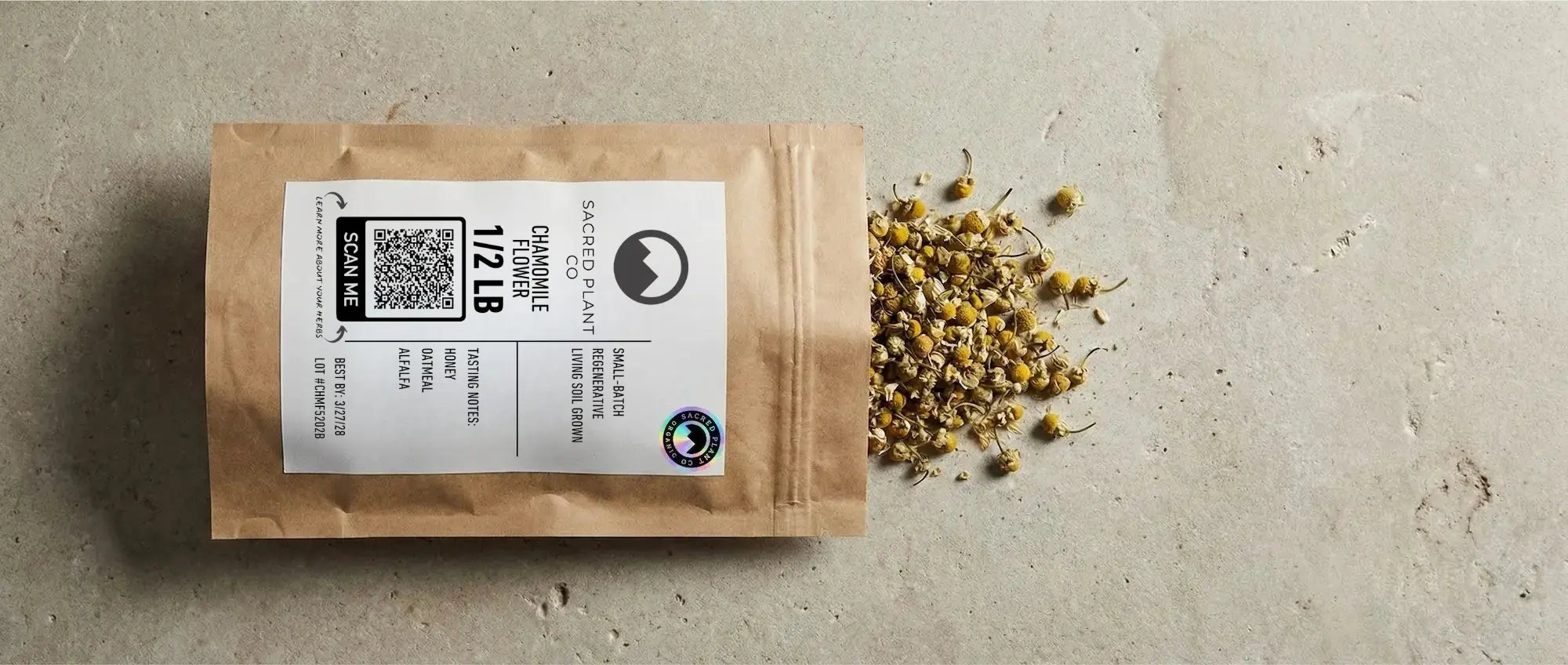 A brown paper bag of Sacred Plant Co's small-batch, regenerative, living soil grown Chamomile Flower, 1/2 LB, with dried flowers spilling onto a concrete surface.