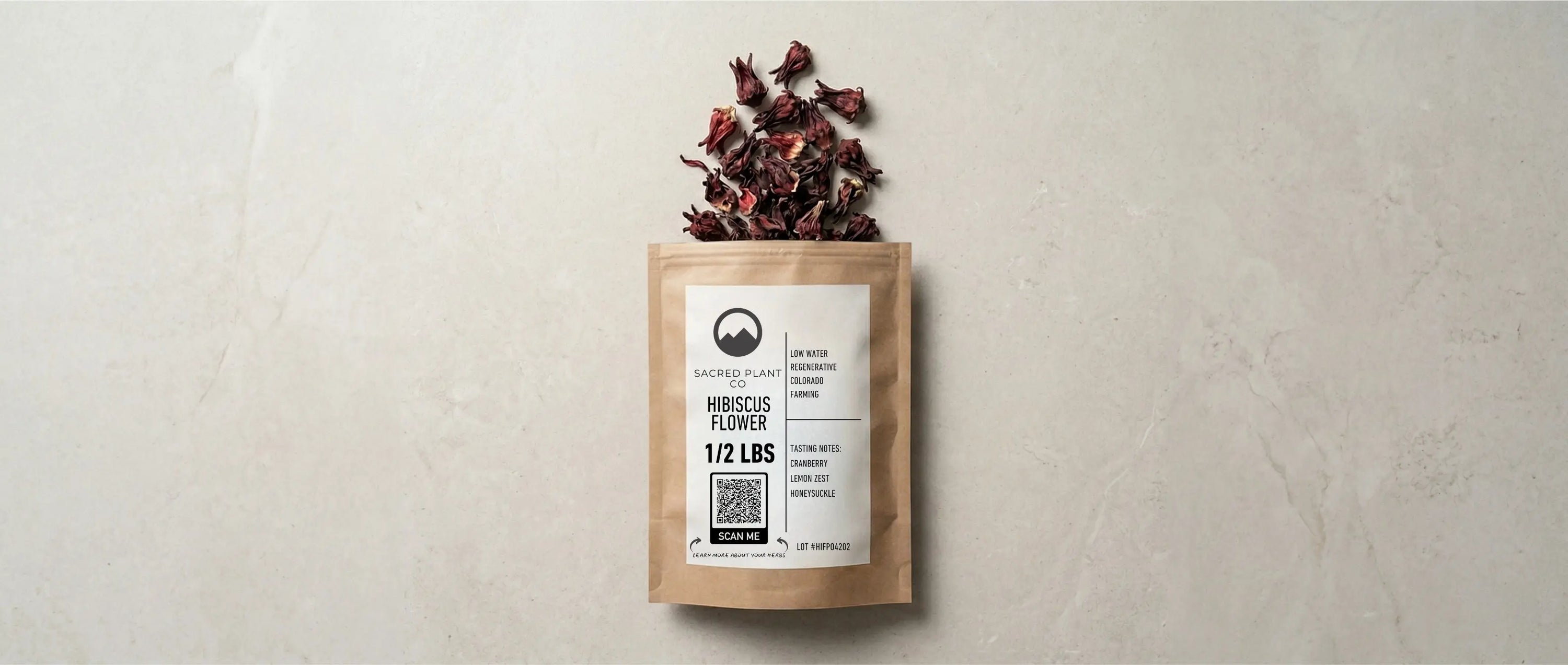 Top-down layflat view of a Sacred Plant Co half-pound kraft paper bag of dried hibiscus flowers, featuring a branded product label, resting on a light textured stone surface with loose red hibiscus calyxes placed near the top opening.