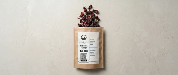 Top-down layflat view of a Sacred Plant Co half-pound kraft paper bag of dried hibiscus flowers, featuring a branded product label, resting on a light textured stone surface with loose red hibiscus calyxes placed near the top opening.