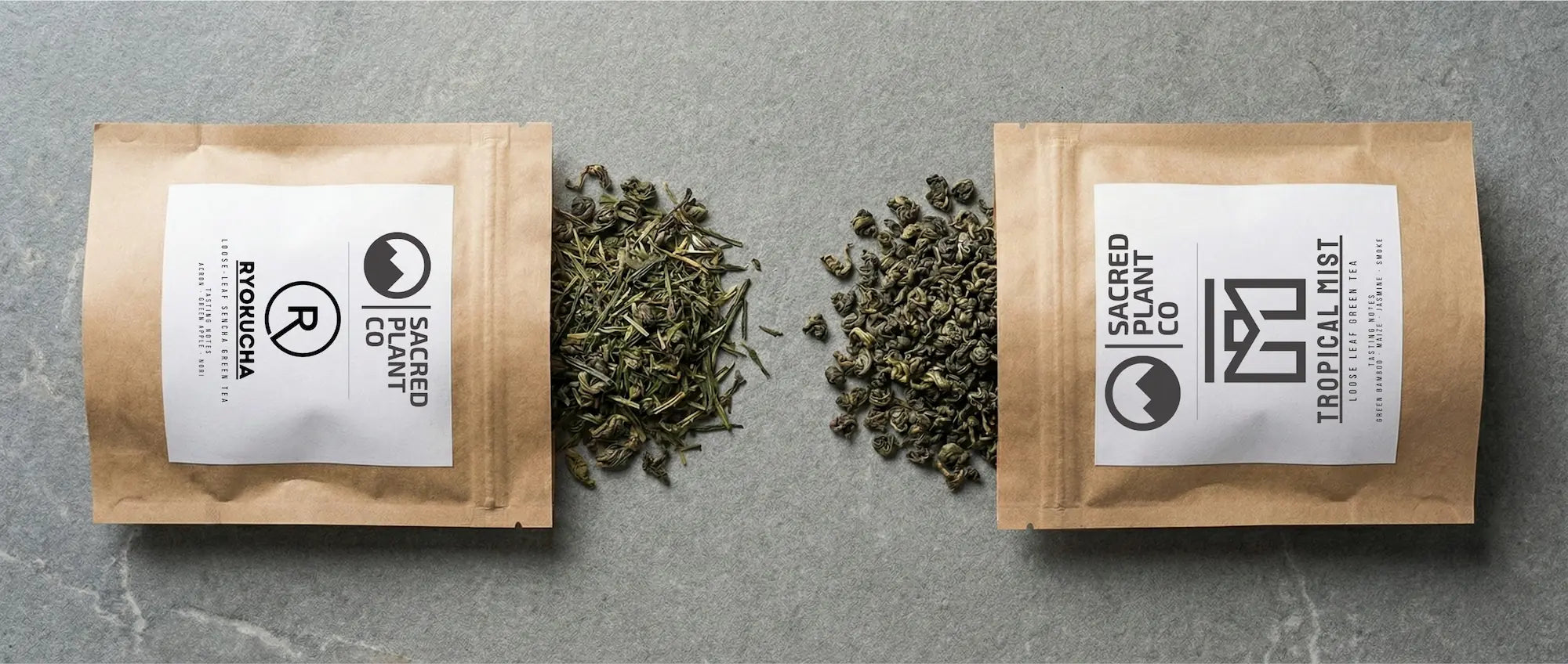 Two kraft paper bags of Sacred Plant Co loose leaf green tea lying flat on a stone surface, featuring Ryokucha Sencha with needle-like leaves on the left and Tropical Mist Chun Mei with curled leaves on the right.
