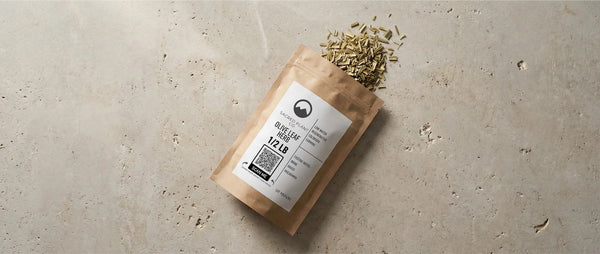 Sacred Plant Co organic olive leaf herb packaging spilling dried cut and sifted leaves onto a textured stone surface, displaying label with "Olive Leaf Herb 1/2 LB" text.