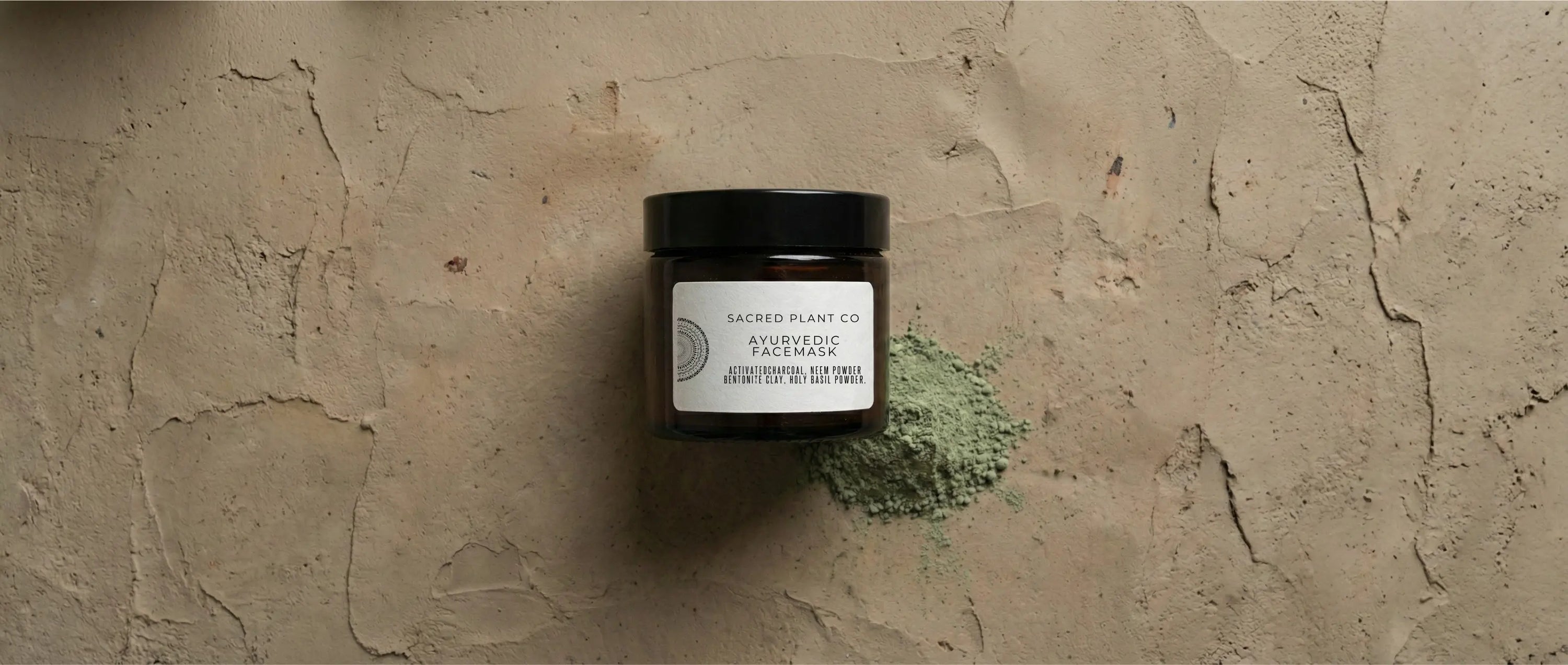 Top-down view of Sacred Plant Co Ayurvedic Face Mask jar beside a pile of fine, olive-green neem and bentonite clay powder on a textured earth-tone surface.