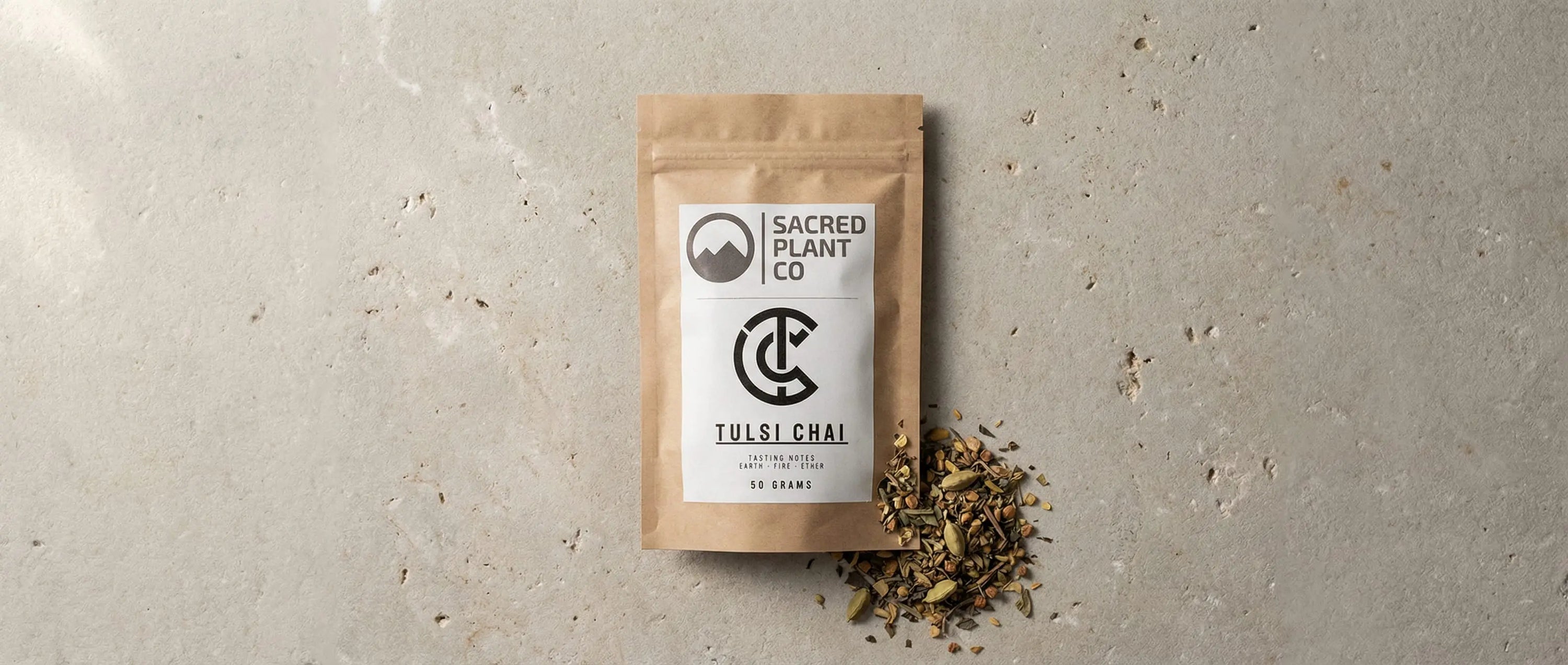 Top-down view of Sacred Plant Co Tulsi Chai package on a stone surface, with loose spices including green cardamom pods, ginger, and tea leaves spilling from the pouch.